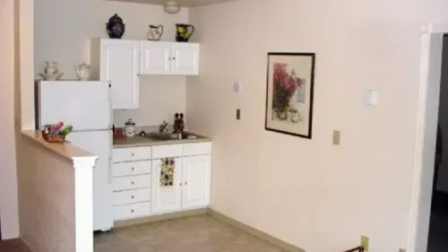 A small apartment kitchen with a white refrigerator, white cabinets, a sink, and a framed picture on the wall